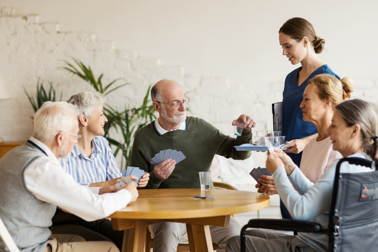 Group of seniors playing cards while nurse gives them cups medication