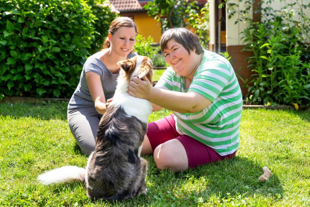 Caregiver and senior sitting down, petting a dog in the grass